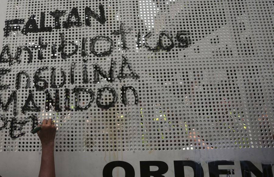 Un manifestante escribe sobre la barricada policiaca protegiendo el ministro de salud la lista de medicamentos que hacen falta para atender diversas enfermedades y que han provocado un éxodo de venezolanos a otros países vecinos en búsqueda de atención médida. Foto AP/Fernando Llano